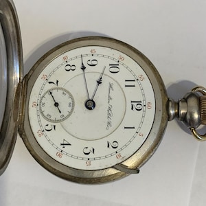 Op de afbeelding: Antieke zakhorloge met een witte wijzerplaat, zwarte cijfers en rode accenten. Het horloge heeft een subwijzerplaat en de inscriptie "Hamilton Watch Co." De zilverkleurige kast is open.