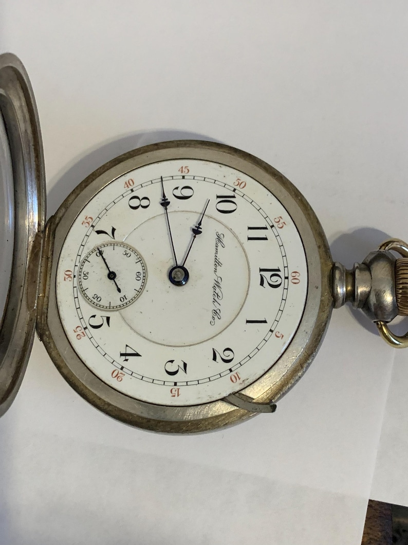 Op de afbeelding: Antieke zakhorloge met een witte wijzerplaat, zwarte cijfers en rode accenten. Het horloge heeft een subwijzerplaat en de inscriptie "Hamilton Watch Co." De zilverkleurige kast is open.
