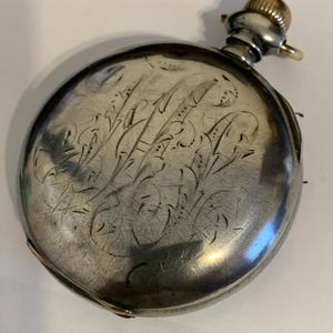 Op de afbeelding: Een zilveren zakhorloge met een gegraveerd bloemmotief op de voorkant. Het horloge heeft een goudkleurige kroon en een kleine lus voor een ketting. Het ronde horloge heeft een licht versleten uiterlijk.