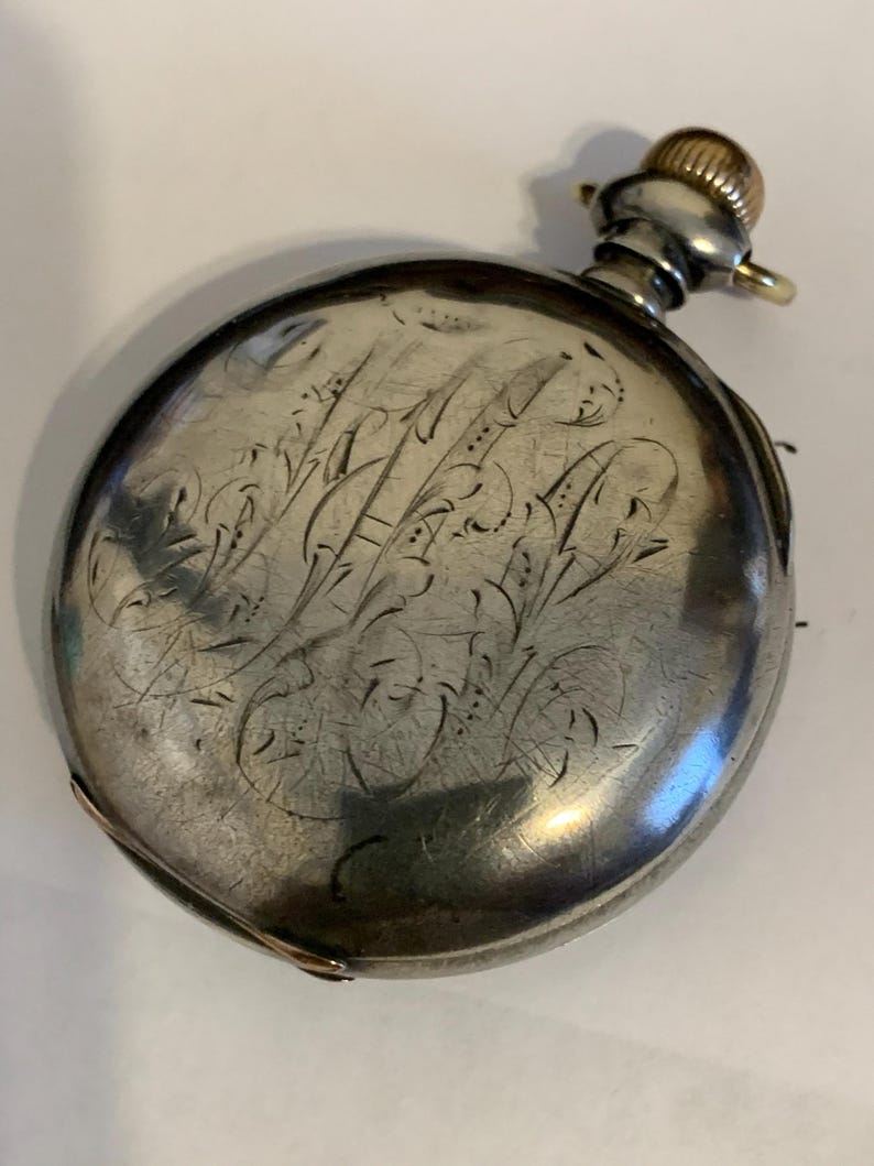 Op de afbeelding: Een zilveren zakhorloge met een gegraveerd bloemmotief op de voorkant. Het horloge heeft een goudkleurige kroon en een kleine lus voor een ketting. Het ronde horloge heeft een licht versleten uiterlijk.