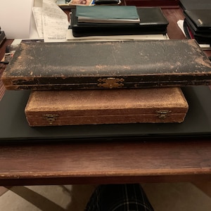 May include: Two vintage wooden boxes with brass clasps. The top box is black and the bottom box is brown.