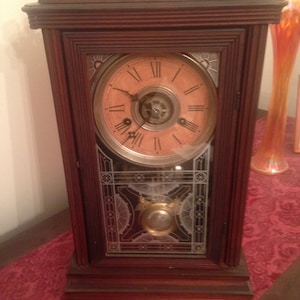 May include: A vintage wooden clock with a decorative glass face and a Roman numeral clock face. The clock has a dark wood frame and a decorative top.