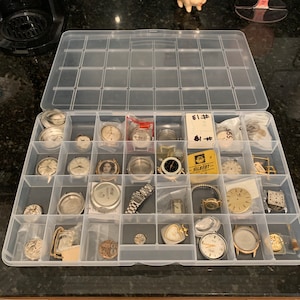 May include: A clear plastic storage box filled with various watch parts and watch faces. The box has 24 compartments and is filled with watch parts, including watch faces, watch hands, watch movements, and watch straps.