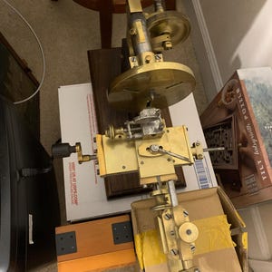 Puede incluir: Un instrumento antiguo de latón y metal con engranajes, palancas y una placa circular. El instrumento está montado sobre una base de madera y parece ser una herramienta de precisión o parte de una máquina más grande. El diseño sugiere que podría ser un dispositivo científico o industrial.