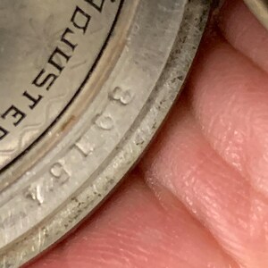 Op de afbeelding: Close-up van een zilveren horlogekast met het woord "ADJUSTED" en het nummer "30154" zichtbaar. De horlogekast heeft een getextureerd, patroonontwerp. De afbeelding is enigszins onscherp.