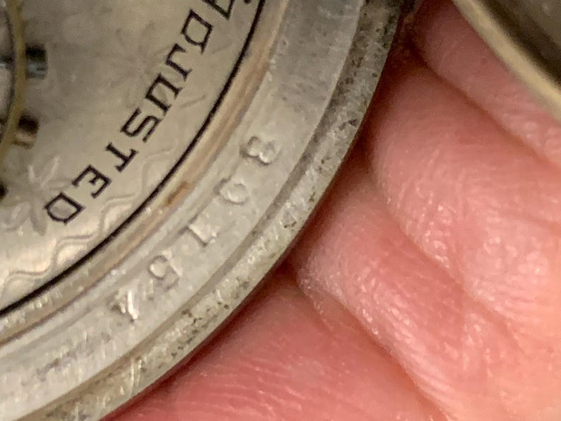 Op de afbeelding: Close-up van een zilveren horlogekast met het woord "ADJUSTED" en het nummer "30154" zichtbaar. De horlogekast heeft een getextureerd, patroonontwerp. De afbeelding is enigszins onscherp.