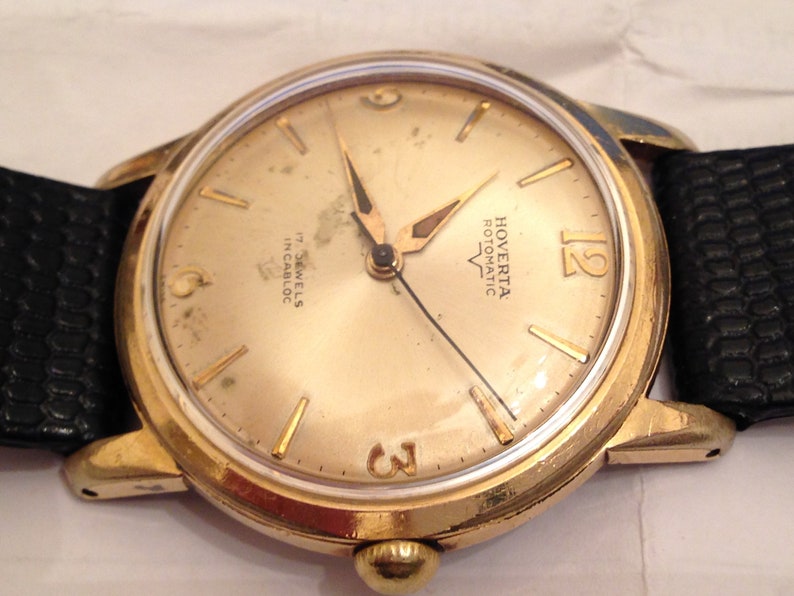 Puede incluir: Un reloj de pulsera dorado con una esfera blanca y agujas negras. La esfera del reloj tiene marcadores de hora en n&uacute;meros romanos negros y el texto "Hoverta Rotonatic 17 Jewels Incabloc" impreso en &eacute;l.