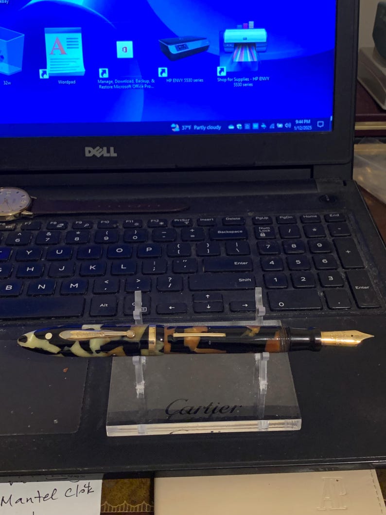 May include: A black, gold, and white patterned fountain pen with a gold nib resting on a black and white stand with the word "Cartier" printed on it.