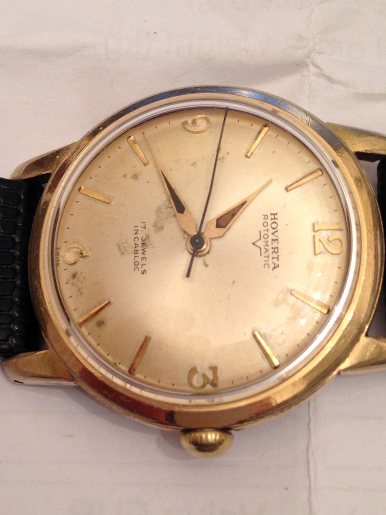 Puede incluir: Un reloj de pulsera dorado con una esfera blanca y agujas negras. La esfera del reloj tiene marcadores de hora en n&uacute;meros romanos negros y una aguja de segundos negra. La esfera del reloj tambi&eacute;n tiene el texto "Hoverta Automatic 17 Jewels Incabloc" impreso en ella.