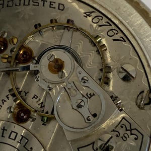 Op de afbeelding: Close-up van het mechanisme van een vintage zakhorloge. De zilverkleurige wijzerplaat toont ingewikkelde tandwielen, juwelen en het woord "ADJUSTED". Het serienummer "46767" is zichtbaar. Het horloge heeft een gedetailleerd, sierlijk ontwerp.