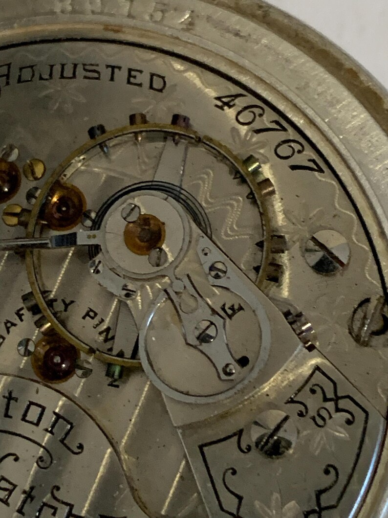 Op de afbeelding: Close-up van het mechanisme van een vintage zakhorloge. De zilverkleurige wijzerplaat toont ingewikkelde tandwielen, juwelen en het woord "ADJUSTED". Het serienummer "46767" is zichtbaar. Het horloge heeft een gedetailleerd, sierlijk ontwerp.