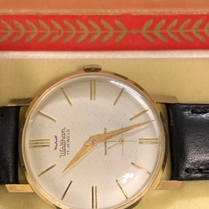 Op de afbeelding: Een goudkleurige Waltham-horloge met een witte wijzerplaat en een zwarte leren band. De wijzerplaat van het horloge heeft zwarte Romeinse cijfers als uurmarkeringen en een gouden secondewijzer. Het horloge is gelabeld met "Waltham 17 Jewels" en "Shockproofed".