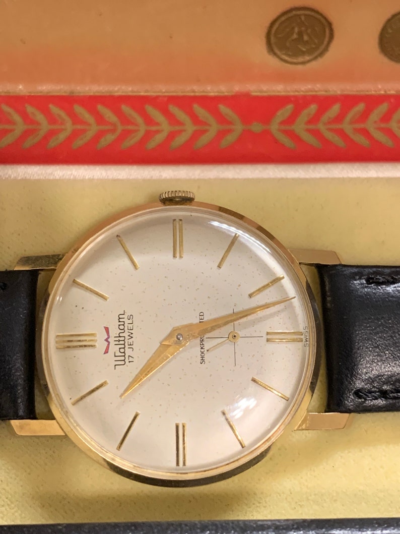 Op de afbeelding: Een goudkleurige Waltham-horloge met een witte wijzerplaat en een zwarte leren band. De wijzerplaat van het horloge heeft zwarte Romeinse cijfers als uurmarkeringen en een gouden secondewijzer. Het horloge is gelabeld met "Waltham 17 Jewels" en "Shockproofed".