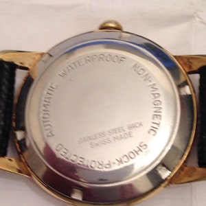 Puede incluir: La parte posterior de un reloj de pulsera dorado con una correa de cuero negro. La esfera del reloj est&aacute; marcada con "Automatic Waterproof Non-Magnetic Shock-Protected Stainless Steel Back Swiss Made".