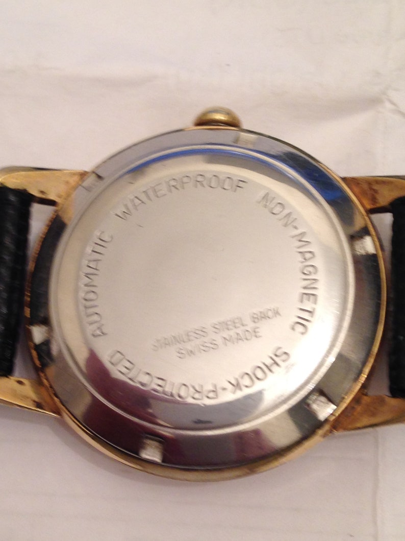 Puede incluir: La parte posterior de un reloj de pulsera dorado con una correa de cuero negro. La esfera del reloj est&aacute; marcada con "Automatic Waterproof Non-Magnetic Shock-Protected Stainless Steel Back Swiss Made".