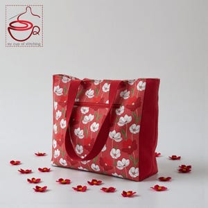 May include: A red tote bag featuring a floral design of white and red flowers. The bag has red handles and a red zip. Small red flower accents are scattered around the base. The bag is likely made of fabric.