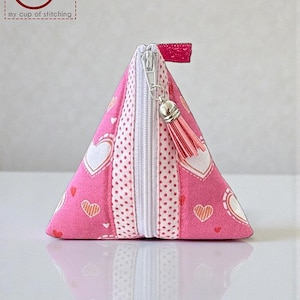 May include: A pink triangular pouch with a white zipper and a tassel. The pouch is decorated with white hearts and red heart outlines on a pink background. The pouch has a small pink loop on the top.
