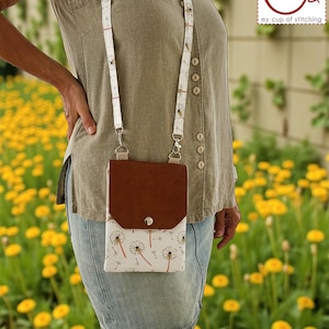 May include: A brown and white crossbody bag with a dandelion pattern, worn by a person. The bag has a brown flap and a white body with a dandelion design. The strap is white with a matching dandelion pattern. The bag is worn over a green top and denim skirt.