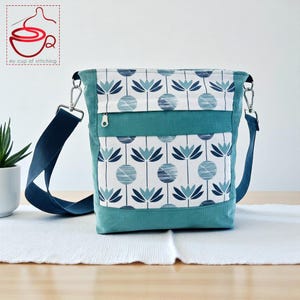 Puede incluir: Bolso bandolera de pana color turquesa con estampado floral. El bolso presenta un fondo blanco con diseños de flores azules. Tiene una correa ajustable azul marino y un pequeño bolsillo con cremallera. El bolso está sobre una superficie blanca.