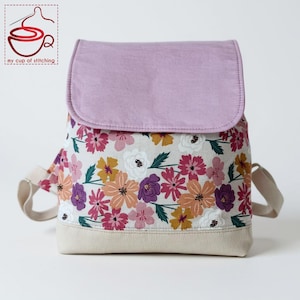 Op de afbeelding: Een bloemenrugzak met een lichtpaarse flap en een crèmekleurige basis. De rugzak heeft een patroon van roze, oranje, gele en paarse bloemen op een lichte achtergrond. De bandjes zijn crèmekleurig en de tas heeft de tekst "my cup of stitching".