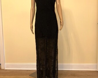 May include: A black formal gown with spaghetti straps. The bodice is embellished with small, dark beads. The skirt features a sheer overlay with a floral pattern. The dress is displayed on a mannequin.