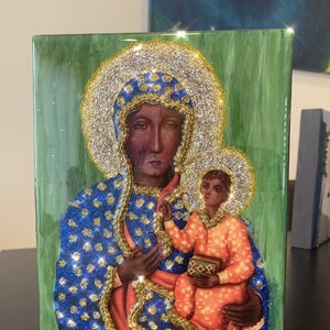 May include: A religious icon featuring a dark-skinned Madonna and child, adorned with gold and glitter accents. The Madonna wears a blue robe with gold stars, and the child holds a small box. The background is a textured green.
