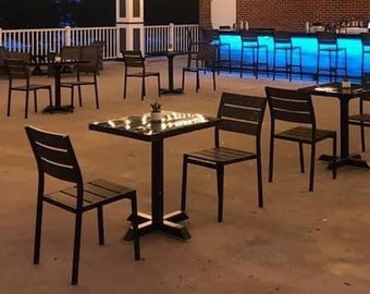 May include: An outdoor patio with a bar and tables. The bar has a blue lit countertop and black bar stools. The tables are made of wood and have black chairs. The patio is decorated with string lights.
