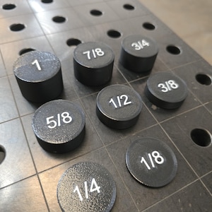 May include: A collection of black metal round plugs with white numbers and fractions, including 1, 7/8, 3/4, 1/2, 5/8, 3/8, 1/4, and 1/8. The plugs are arranged on a metal surface with evenly spaced holes.