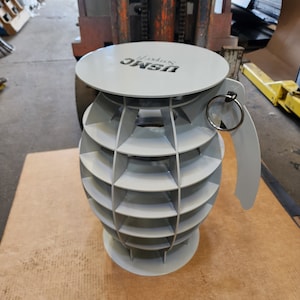 May include: A white metal grenade-shaped container with a round top and a ring handle. The top has the words "USMC" and "Semper Fidelis" cut out.