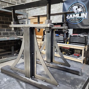 May include: Two unfinished metal table legs with adjustable height features. Each leg has a rectangular base, a central support, and a triangular brace. The top plate is designed to hold a tabletop. The legs are designed for welding and fabrication.