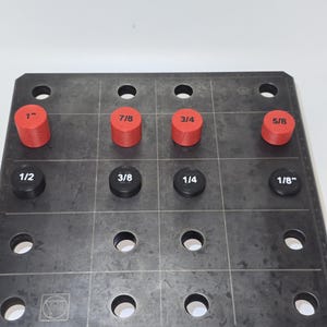May include: A dark gray metal work surface with a grid pattern, featuring red and black cylindrical measuring tools labeled 1", 7/8, 3/4, 5/8, 1/2, 3/8, 1/4, and 1/8". The tools are arranged in a grid, with circular holes around them.