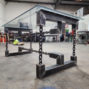 May include: A glass-top table with a unique industrial design. The table base is made of metal and features a series of connected chains. The base is marked with the text "3481KA-B7U".