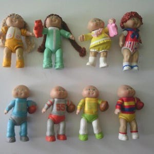 May include: A collection of eight small plastic dolls with round faces and various outfits. The top row features dolls with blonde hair, brown pigtails, yellow clothing, and red hair. The bottom row shows dolls in blue, grey, yellow, and striped shirts.