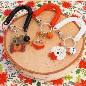 May include: Three colorful retractable keychains with festive animal charms. The charms are a dog wearing glasses, a cat wearing a Santa hat, and a poodle wearing a red scarf. Each keychain has a silver key ring and a bell.