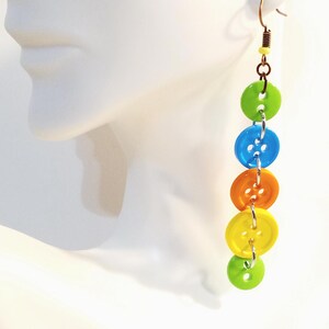 May include: A dangle earring featuring five circular buttons in a vertical arrangement, connected by small silver-toned metal jump rings. The buttons are colored lime green, bright blue, orange, yellow, and a smaller lime green button at the base.