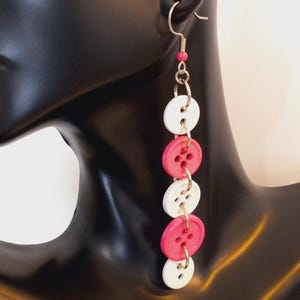 May include: A long drop earring featuring a vertical arrangement of five alternating white and hot pink circular buttons connected by silver-toned metal jump rings, suspended from a silver-toned hook with a small pink bead at the top.
