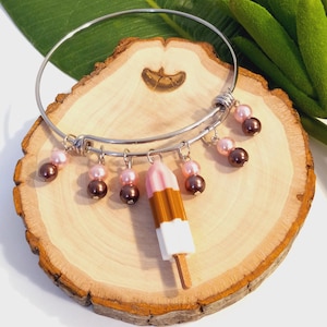 May include: A silver-colored bangle bracelet with pink and brown pearl beads and a popsicle charm. The popsicle charm has a white, pink, and brown design. The bracelet is displayed on a wooden slice.
