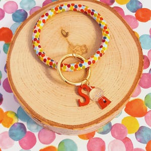May include: A colorful polka dot silicone key ring with a gold key chain and a red letter "S" charm with a clear gumball machine charm.