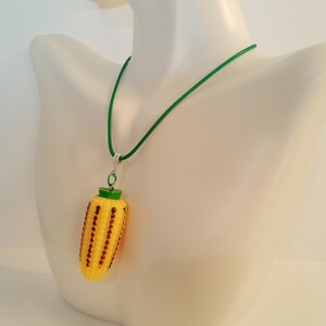 May include: A green cord necklace with a yellow corn on the cob pendant. The corn is decorated with brown and green details.