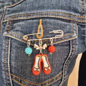 May include: A silver-toned safety pin brooch featuring three dangling charms: a turquoise blue glitter bead, a central silver bow, and a red glitter bead. Below the bow hangs a pair of miniature red shoes with silver-toned accents and detailing.