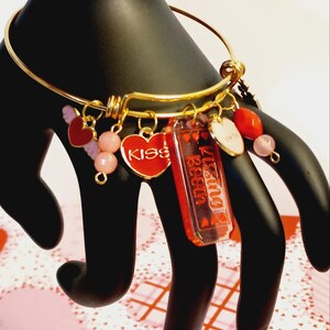 May include: Gold-tone bangle bracelet with multiple charms. Charms include a red heart with "KISS" text, a red rectangle with "Kissing Begin" text, a heart with wings, pink beads, and other heart-shaped charms. The bracelet is displayed on a black hand.