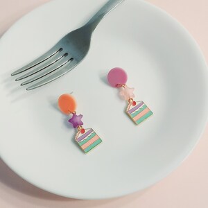 May include: A pair of dangle earrings featuring a layered cake charm with a pink cherry on top, a purple star, and a pink and orange circle. The earrings are on a white plate.