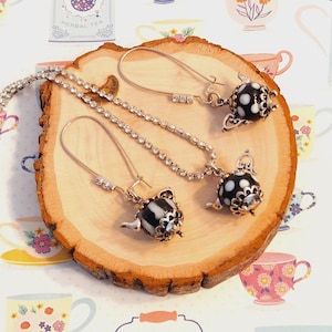 May include: A set of black and white teapot earrings and a matching necklace with a rhinestone chain. The jewelry is displayed on a wooden slice.