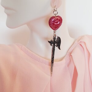 May include: A pair of dangle earrings with a silver axe charm and a red heart-shaped bead. The earrings are hanging from a silver chain.