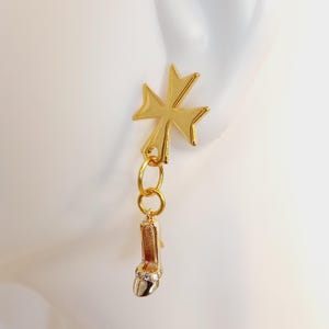 May include: Gold-colored earring featuring a Maltese cross design at the top, connected by small gold rings to a high-heeled shoe charm. The shoe charm has a rhinestone-studded toe and a slender heel. The earring is displayed against a white background.