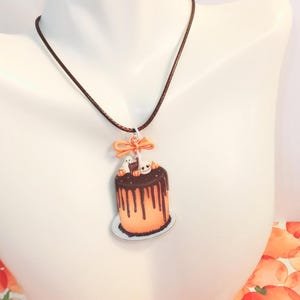 May include: A necklace featuring a pendant shaped like an orange cake with dark brown chocolate ganache drips. The cake is topped with a small white ghost, a skull, and tiny orange pumpkins. An orange bow sits above the cake on a brown cord.