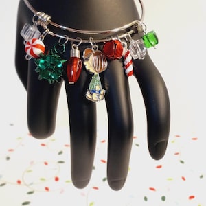 May include: A silver bangle bracelet adorned with Christmas-themed charms. Charms include a red and white peppermint candy, a green bow, a red light bulb, a gingerbread person, a red bell, a candy cane, and clear and green crystal-like charms. The bracelet is displayed on a black hand.