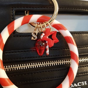 May include: A black handbag with a gold zipper and a red and white striped bracelet. The bracelet has gold charms, including a letter "S", a Christmas light, a candy cane, and a star.
