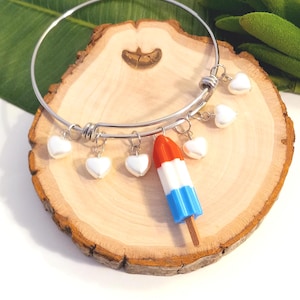 May include: A silver bangle bracelet with white heart charms and a red, white, and blue popsicle charm. The bracelet is displayed on a wooden slice with a white background. The popsicle charm is a patriotic design.