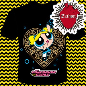 Op de afbeelding: Zwart T-shirt met een gouden hartontwerp met een cartoonfiguur met blond haar en blauwe ogen. Het personage draagt een blauwe en witte jurk en houdt haar hand omhoog. De tekst "The Powerpuff Girls" is onder het hartontwerp gedrukt.